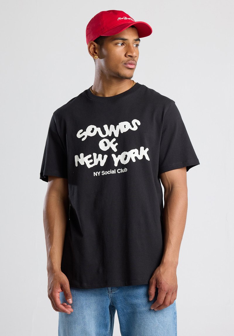 Black cotton t-shirt features white "SOUNDS OF NEW YORK" text. Paired with a red baseball cap. Simple design, casual fit.