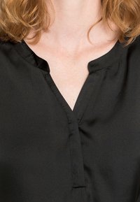 Svart blus med ett slätt, glänsande tyg; har en V-halsringning och subtila sömmar längs kragen. Enkelt och elegant design.