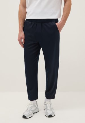 Homme portant un pantalon de jogging bleu marine avec des poignets élastiques et la main dans la poche, assorti de baskets blanches et de chaussettes blanches.