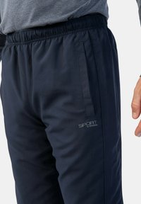 Navyblaue Sporthosen mit einem Gummizugbund, seitlichen Taschen und einem grauen "SPORT by STOOKER"-Logo auf dem Oberschenkel. Glatte Textur, moderner Schnitt.