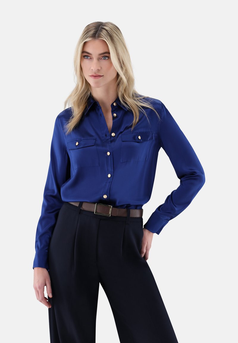 Royal blue satin blouse with long sleeves, two chest pockets, and gold button accents, paired with high-waisted black trousers and a brown belt.