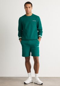 Grøn sweatshirt med kontrasterende stribede manchetter og matchende shorts, med broderet "Calvin Klein" branding. Båret med hvide sneakers.