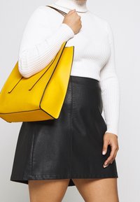 Borsa a tracolla gialla in pelle con dettagli neri, abbinata a un dolcevita bianco a coste e a una mini gonna nera in ecopelle.