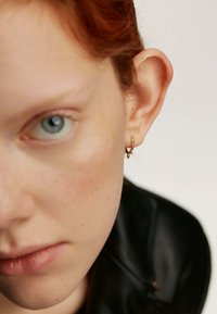 Pendientes de aro de oro con pequeños acentos de corazón colgantes. Primer plano de la oreja de un modelo, mostrando la joyería contra un tono de piel claro y una textura suave.