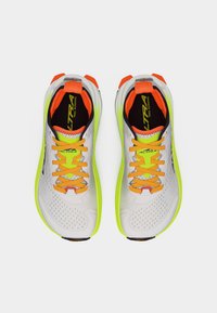 Ζευγάρι λευκά, νέον κίτρινα και πορτοκαλί παπούτσια για τρέξιμο σε μονοπάτια με πορτοκαλί κορδόνια, που φαίνονται από πάνω σε απλό λευκό φόντο.