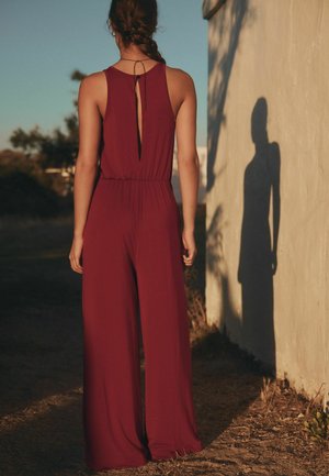 Frau mit geflochtenem Haar steht mit dem Rücken zur Wand, trägt einen ärmellosen, bordeauxroten Overall mit weiten Beinen und wirft einen Schatten an die Wand.