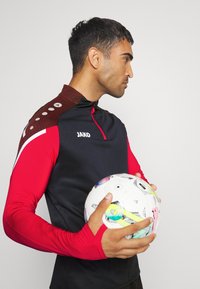 Langärmliges Sportoberteil in Marineblau, rote Ärmel und gemusterte kastanienbraune Schulter. Das Modell hält einen mehrfarbigen Fußball mit geometrischen Mustern.