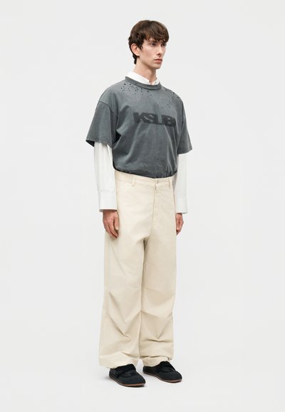 Jeune homme debout, portant une chemise grise à manches courtes avec un texte par-dessus une chemise blanche à manches longues, un pantalon beige à jambes larges et des chaussures noires.