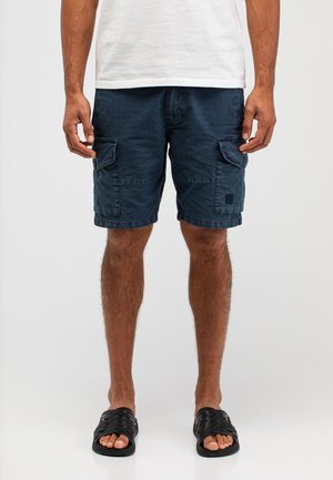 Man die donkerblauwe cargoshorts, een wit T-shirt en zwarte slip-on sandalen draagt, staand tegen een effen lichte achtergrond.