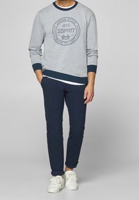 Sweat-shirt gris avec poignets et col bleu foncé, arborant un logo Esprit central. Associé à un pantalon de jogging bleu marine et des baskets blanches.