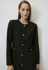Black, long-sleeve dress with a round neck, featuring gold buttons and structured pockets, made of textured fabric. Smooth finish overall.
