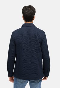 Marineblau gesteppte Jacke mit Köperstruktur, Kragen, langen Ärmeln und geradem Schnitt. Getragen mit blauen Jeans.