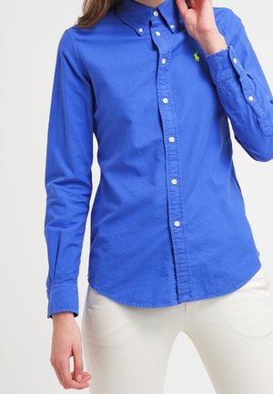 Frau trägt ein eng anliegendes, leuchtend blaues Hemd mit Knöpfen und einem kleinen grünen Logo auf der Brust sowie weiße Hosen vor hellem Hintergrund.