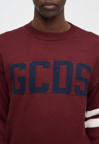 Burgunderfarget strikkegenser med en stor marineblå "GCDS"-logo, ribbet hals og hvite og marineblå stripete detaljer på ermene.