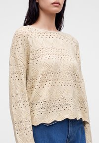 Femme portant un pull en maille beige avec des motifs en dentelle élaborés et un ourlet festonné, associé à un jean bleu sur un fond uni.
