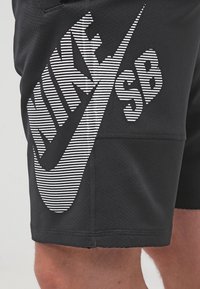 Černé sporty kraťasy z texturované látky, s výrazným bílým pruhovaným logem Nike SB na levé straně.
