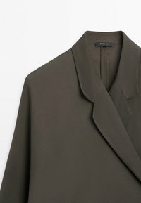 Blazer vert olive foncé avec col à revers cranté et étiquette de marque visible à l'intérieur du col.