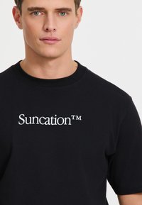 Juodas medvilninis marškinėliai su balta logo „Suncation™“ priekyje. Trumpomis rankovėmis, apvaliu kaklu ir lygiu paviršiumi.