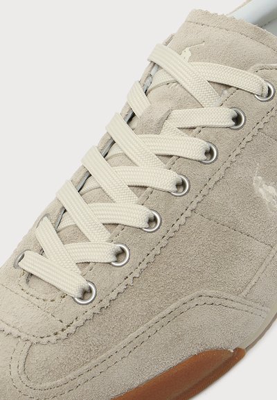 Gros plan sur une basket en daim beige avec des lacets crème, des œillets métalliques et une semelle en caoutchouc marron sur fond blanc uni.