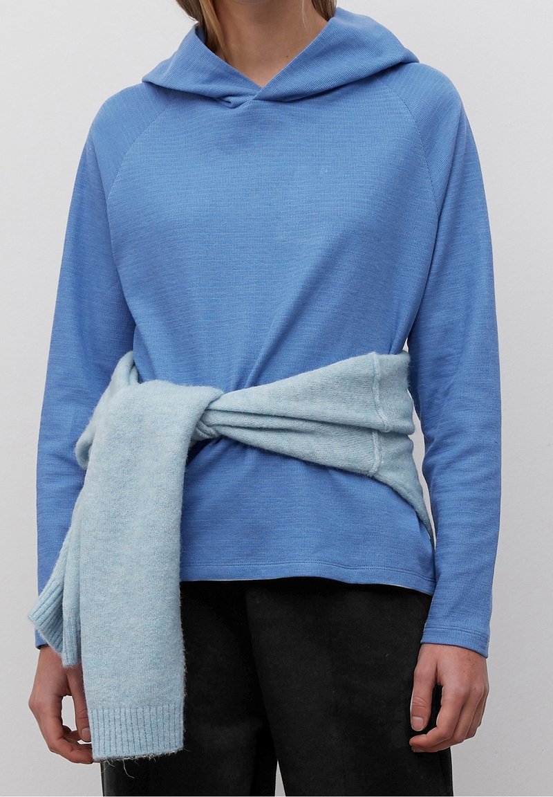 Blå hoodie med texturerad yta och huva med dragsko. En ljusblå, mjuk halsduk är knuten runt midjan. Svarta byxor bärs nedanför.