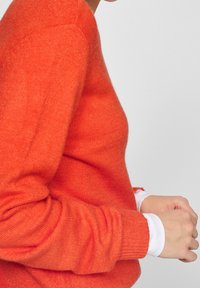 Pull en maille orange avec une texture douce et des manches longues, doté d'un poignet côtelé et d'une silhouette ajustée, porté sur une chemise blanche.