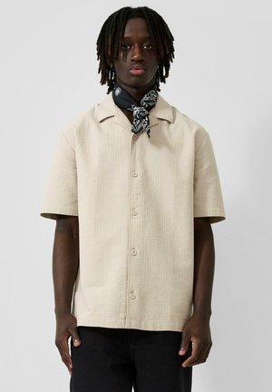 Mann mit mittel langen Dreadlocks trägt beige kurzärmliges Hemd mit Knöpfen und ein schwarzes Bandana, das um den Hals gebunden ist, und steht vor einfachem Hintergrund.