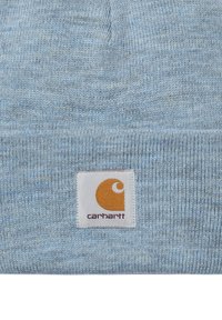 Gorro de punto azul claro con una textura suave y un puño doblado, con una etiqueta rectangular blanca que muestra un logo naranja y el nombre de la marca.