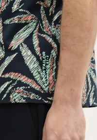 Korte mouw shirt met een marineblauwe achtergrond en groene en koraalkleurige bladenpatronen, en het "TOM TAILOR"-logo in lichtblauw aan de onderkant.