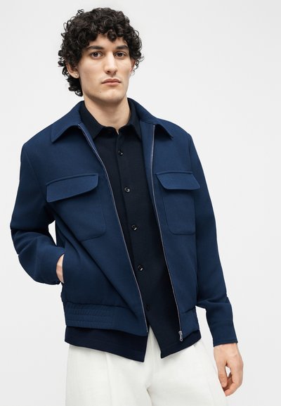 Νέος άνδρας με σγουρά μαλλιά, φορώντας μπλε σκούρο μπουφάν με φερμουάρ πάνω από μαύρο πουκάμισο και λευκό παντελόνι, στέκεται μπροστά σε ένα απλό φωτεινό φόντο.