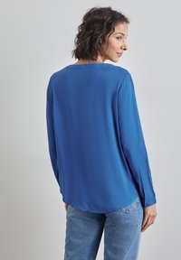 Camicia a maniche lunghe di un blu vivace, con una texture liscia e orlo arrotondato. Dettagliata con polsini abbottonati e una vestibilità rilassata. Abbinata a jeans azzurri.