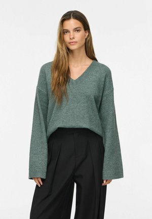 Mujer joven con cabello largo y castaño que lleva un suéter verde holgado de cuello en V y pantalones negros de pierna ancha y cintura alta, de pie frente a un fondo liso.