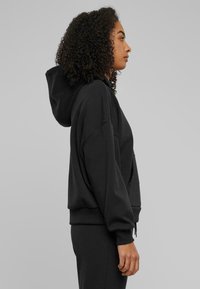 Felpa nera con cappuccio regolabile, vestibilità morbida, polsini larghi e tasca a marsupio. Tessuto morbido e aspetto strutturato. Indossata con pantaloni coordinati.