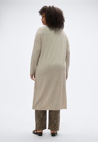 Μπεζ φόρεμα με μακριά μανίκια και χαλαρή εφαρμογή. Συνδυάζεται με παντελόνια καφέ και μαύρα, φαρδιά, με σχέδιο ζιγκ-ζαγκ.