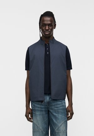 Marineblå, ærmeløs vest med høj krave, frontlynlås og letvægtsstof, båret over en mørk polotrøje og kombineret med blå denimshorts.