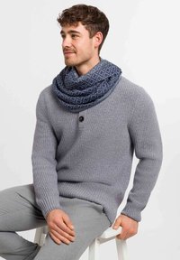 Joven sentado en un taburete blanco, vestido con un suéter de punto gris, pantalones grises y una bufanda infinita de patrón azul, mirando hacia la izquierda.