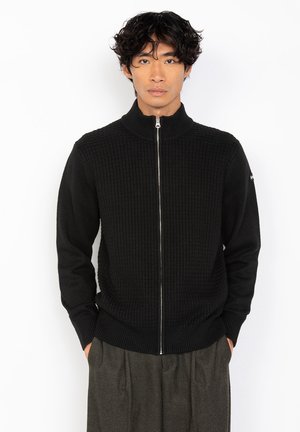Sudadera negra con cremallera y un patrón texturizado en la parte superior del cuerpo, cuello y puños acanalados, y mangas lisas. Presenta una cremallera plateada.