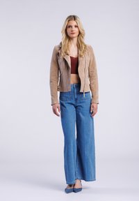 Suède beige motorjack met ritsdetails, gedragen over een bordeauxrood crop top. Losvallende lichtblauwe flared jeans en blauwe flats.