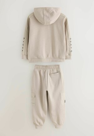 Beżowa bluza z kapturem i pasujące spodnie dresowe. Gładka tkanina, elastyczne mankiety i pas, boczne kieszenie oraz nadruk na rękawie.