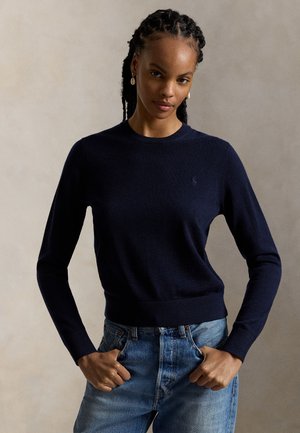 Ung kvinde med fletninger iført navyblå langærmet sweater og blå jeans, stående med hænderne i lommerne mod en ensfarvet baggrund.