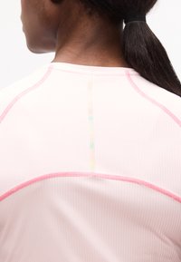 Κοντινό πλάνο του πάνω μέρους της πλάτης ενός ατόμου που φοράει ένα ανοιχτό ροζ πουκάμισο με φωτεινές ροζ λεπτομέρειες στις ραφές και έναν μαύρο λαστιχάκι που συγκρατεί μια ουρά αλόγου.