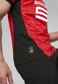 Jersey rojo y negro con un diseño texturizado, con letras blancas y una etiqueta de logo dorada. El material es transpirable y ajustado.