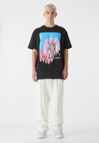 Črna bombažna majica z barvitim grafičnim prikazom abstraktnega cveta na modri podlagi, v kombinaciji s svobodnimi belimi flis hlačami.