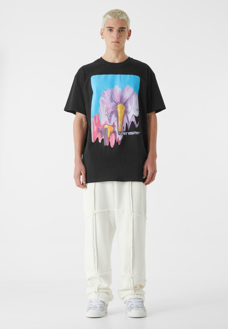 Črna bombažna majica z barvitim grafičnim prikazom abstraktnega cveta na modri podlagi, v kombinaciji s svobodnimi belimi flis hlačami.