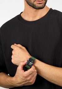 Bracciale in silicone verde con quadrante rettangolare nero che mostra l'ora e delle icone, indossato su un braccio di pelle chiara, sopra una maglietta nera a trama.