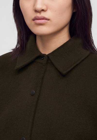 Personne aux cheveux raides et foncés portant un manteau vert olive foncé avec un grand col et des boutons-pression noirs visibles.