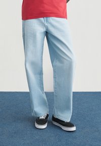 Ανοιχτό μπλε τζιν με ίσια γραμμή και απαλή υφή, συνδυασμένο με μαύρα αθλητικά παπούτσια με λευκές σόλες. Χωρίς εμφανή σχέδια ή μεταλλικά στοιχεία.