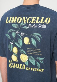 Personne portant une chemise bleu marine avec un motif de citrons jaunes et de feuilles vertes, texte "Limoncello Dolce Vita La Gioia di Vivere" dans le dos.