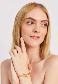 Kožený náramok v svetlohnedej farbe s detailom uzla v zlatej farbe, cylindrické konce a hladký povrch. Ruka modelky jemne sa dotýka jej tváre.