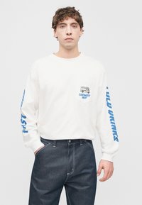 Λευκό μακρυμάνικο μπλουζάκι με μπλε γραφικά στα μανίκια και στην τσέπη. Συνδυάζεται με σκούρο τζιν παντελόνι. Απλός σχεδιασμός, casual εφαρμογή.