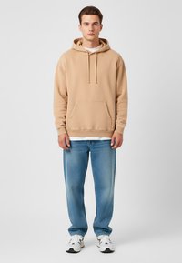 Felpa in cotone beige con tasca frontale e cordino, indossata sopra una camicia bianca. Combinata con jeans azzurri e sneakers bianche.